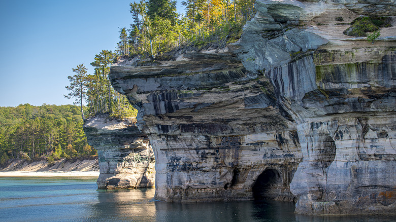 Calm blue waters of Lake Superior lap against the sandstone cliffs of Pictured Rocks National Lakeshore, Munising, Michigan, with pines and early fall foliage above.