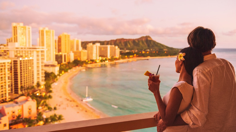 A couple holding a drink and a flower look out over the beach and bay of Honolulu, Hawaii, at sunset