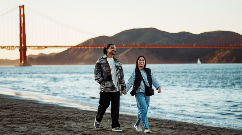 Couple walking hand in hand along the beach below the Golden Gate Bridge in San Francisco, California, at sunset