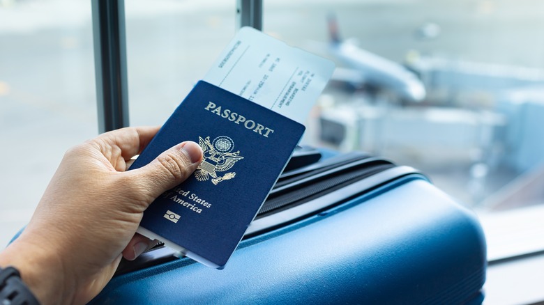 A hand holds out a passport containing a boarding pass, with roll-on luggage in the background.