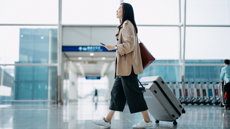 A woman walks through the airport with her luggage, holding her cell phone.