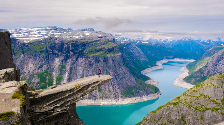 Hiker in red stands on the edge of Norway's dramatic rocky outcrop known as Trolltunga, overlooking a dramatic fjord landscape