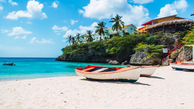 Traditional red and white fishing boat on white sand beach below cliff with colorful homes on the coast of Curaçao