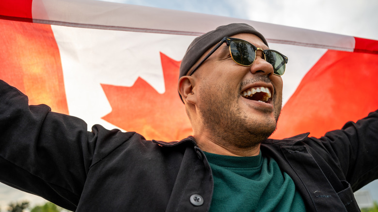 Happy man with black scruff in sunglasses holding a Canadian flag outside and grinning