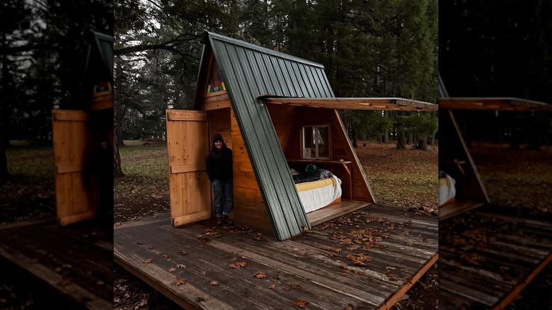 A woman standing in the doorway of a small A-frame cabin in the woods