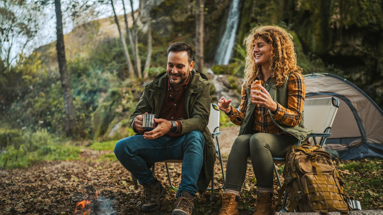 a man and woman sitting on camping chairs by a camp fire with tent in background