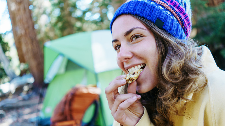 woman wearing a beanie hat eating a granola bar with tent in background