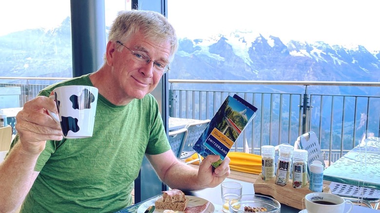 Travel writer Rick Steves sits at a breakfast table in a Swiss Alpine hostel holding a cow-print coffee cup and a travel guidebook