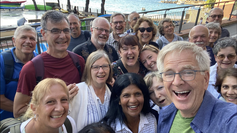 Travel guide Rick Steves smiles during a selfie with a group of smiling people near a waterfront