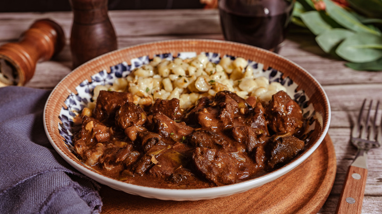 Close-up of steaming beef goulash in a painted dish served with spaetzle dumplings on a wooden table