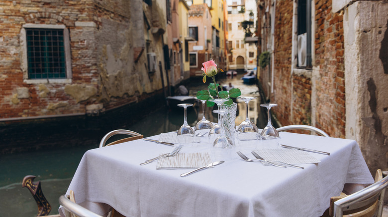 Table set with a pink rose centerpiece, cutlery, and wine glasses, overlooking a narrow canal in Venice, Italy