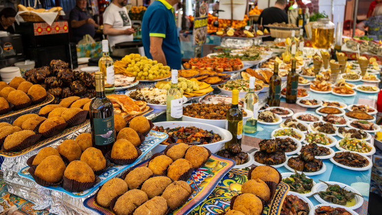 A wide selection of Sicilian street food and local wine on display at Mercato Ballarò, with vendors and customers in the background