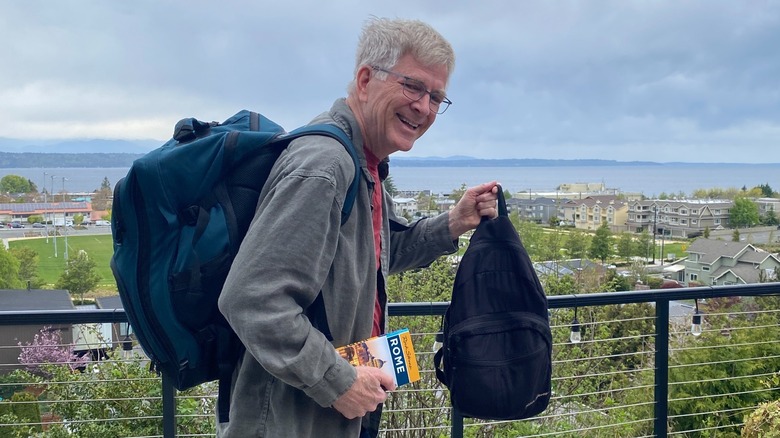 Rick Steves posing in front of a scenic overlook with a large backpack and a small day pack.