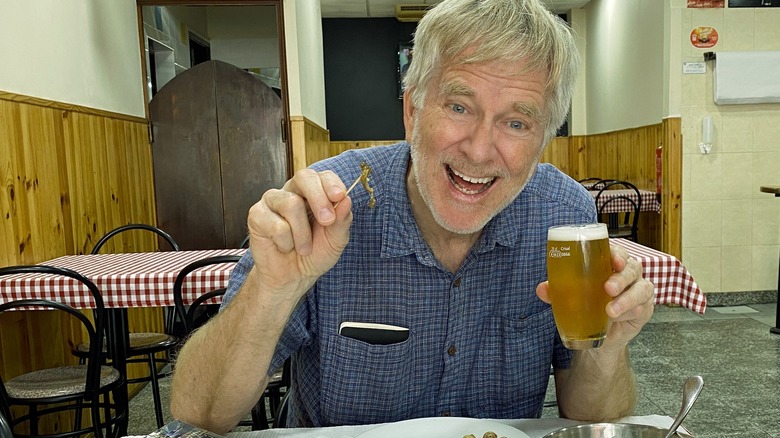 Rick Steves smiling while holding a beer and escargot appetizer at a restaurant table in Portugal.