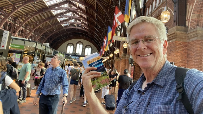 Rick Steves smiling and holding a guidebook inside a brick building