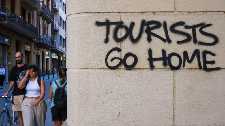 People walking down the street, passing by graffiti on wall reading, "Tourists go home"