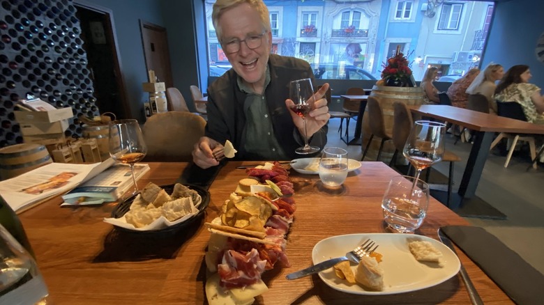 Rick Steves at a restaurant, holding up a glass of wine over a spread of food on a table