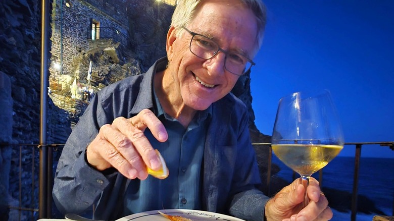 Rick Steves smiling at the camera while holding a lemon over a plate, with a wine glass in the other hand