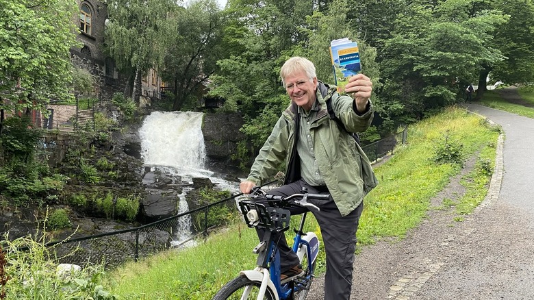 Rick Steves riding a bike in a park in Oslo, holding up his travel guide to Scandinavia