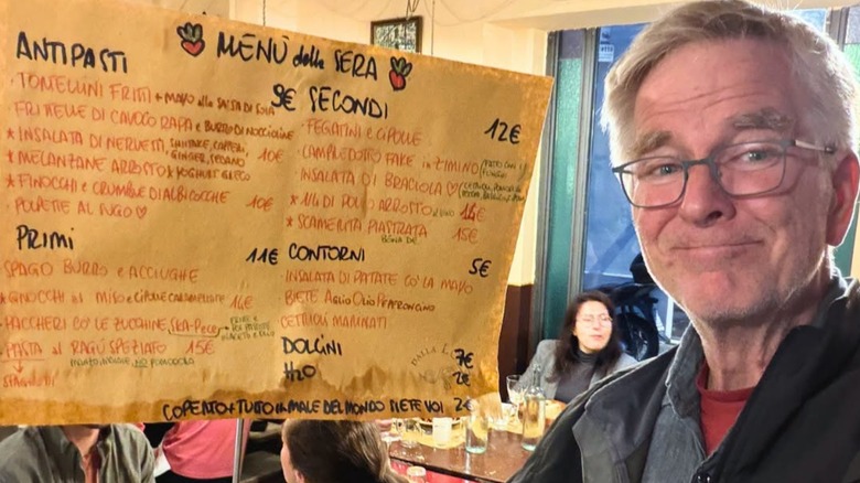 Rick Steves posing next to a handwritten paper menu in a restaurant