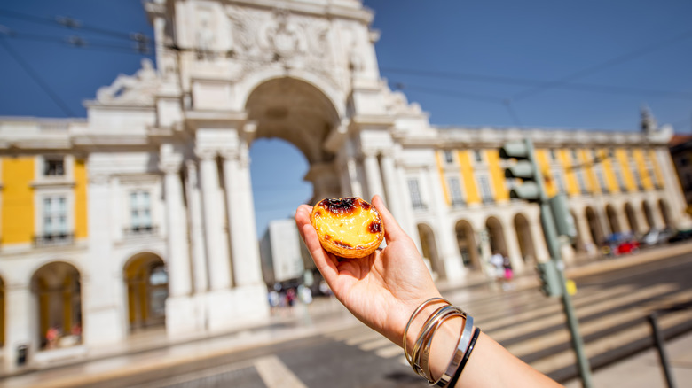 Holding portuguese egg tart pastry called pastel de Nata outdoors on the triumphal arch background in Lisbon