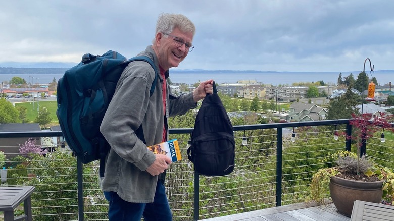 Rick Steves standing on a terrace balcony, holding a backpack and a guidebook