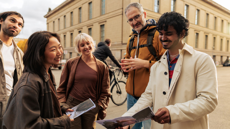 A male tour guide with a backpack talking with a diverse group of tourists while looking at a city map in Berlin, Germany.