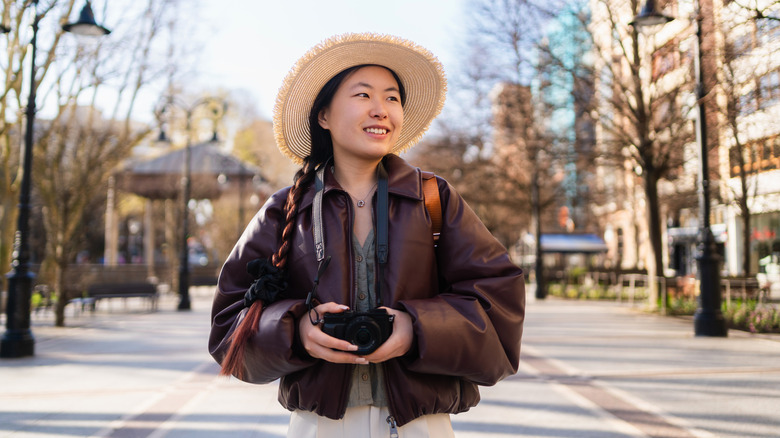 Smiling young Asian woman with braided hair holding a camera while exploring a city park.