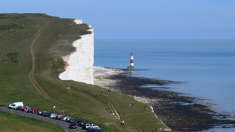 Beachy Head cliff viewed from above, with parked cars and a lighthouse on the coast
