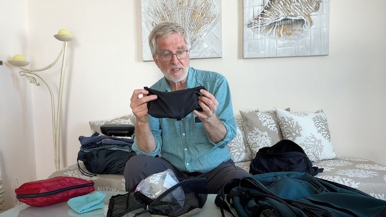 Rick Steves holding a bag, with the contents of his luggage surrounding him on a bed