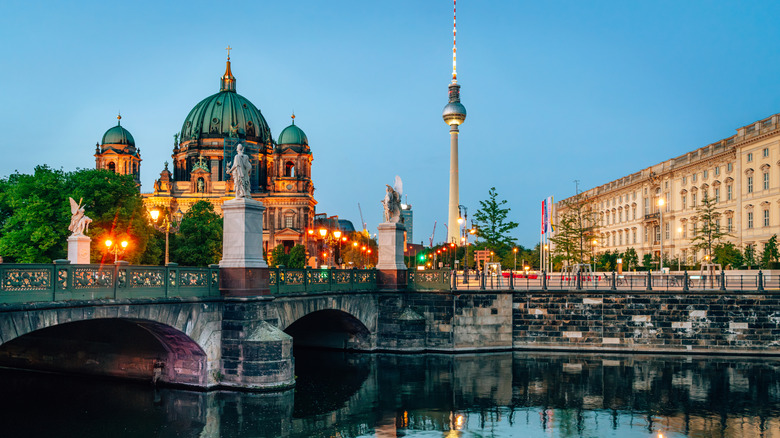 Tranquil scene with Berlin Cathedral, iconic Fernsehturm tower, bridge statues, and evening reflections on water.