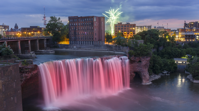 High Falls illuminated at night with fireworks and Rochester skyline in the background.