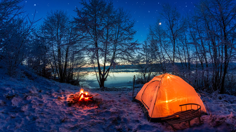 A glowing orange tent and a campfire are pictured with snow on the ground next to a lake at dawn.