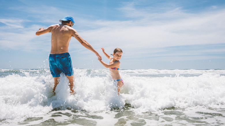 A father and daughter hold hands while jumping breaking waves