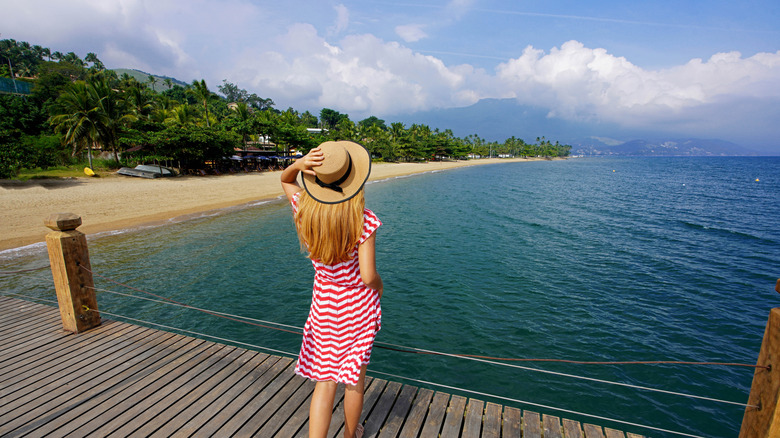A woman wearing a sundress and a hat on a dock, looking at the water near a beach
