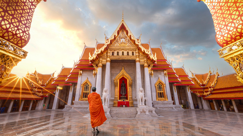 Monk in orange robes stands in courtyard of ornate golden Grand Palace in Bangkok at sunset