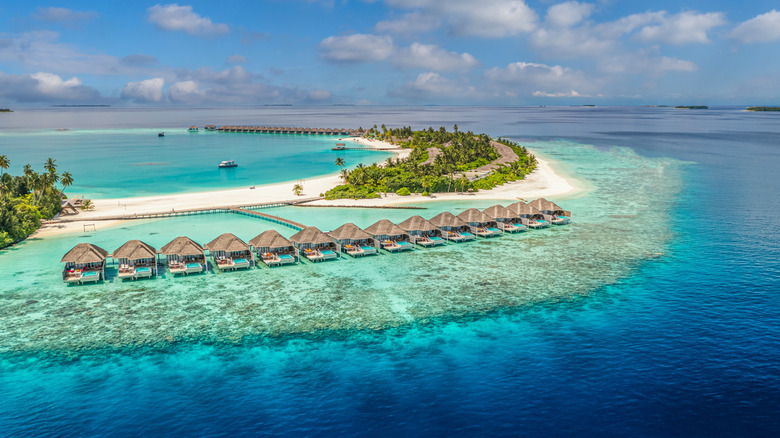 Aerial view of overwater cabanas with thatched roofs on a private beach in the Maldives surrounded by bright blue water and white sand