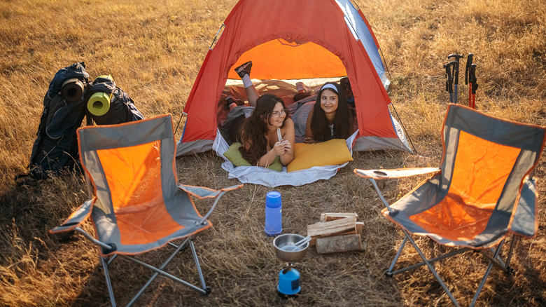 Two young women look out from a tent, with chairs and backpacks nearby