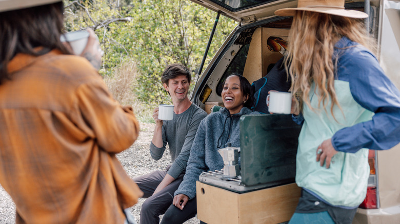 A group of friends sits outside the back of a car with mugs and a camp stove