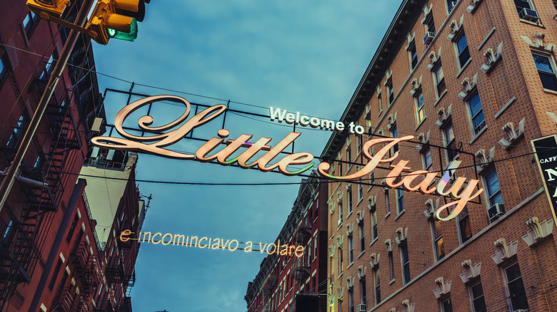 Close-up of "Welcome to Little Italy" sign lit up at dusk strung between two urban brick buildings