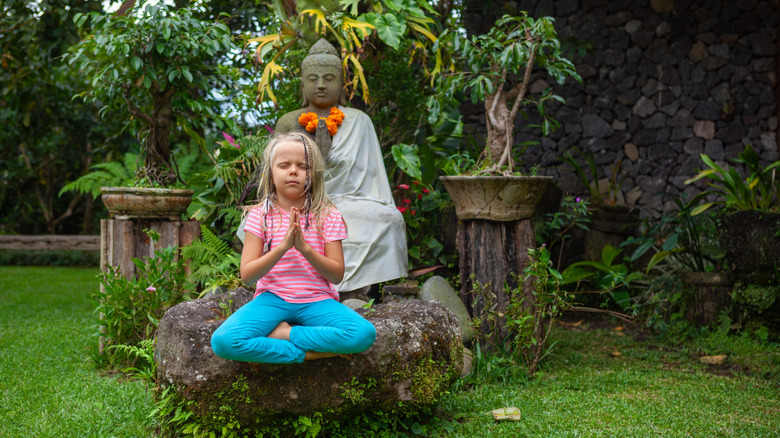 Young blond girl meditates in front of a Buddha statue in a Balinese garden