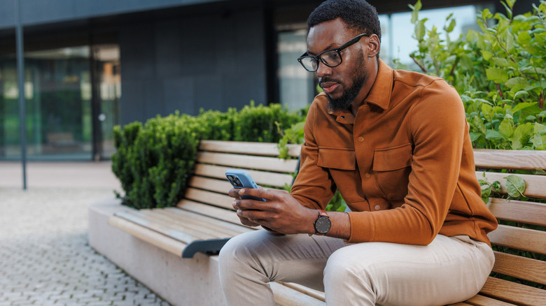 A Black man sits on a bench in an urban area and looks at his phone