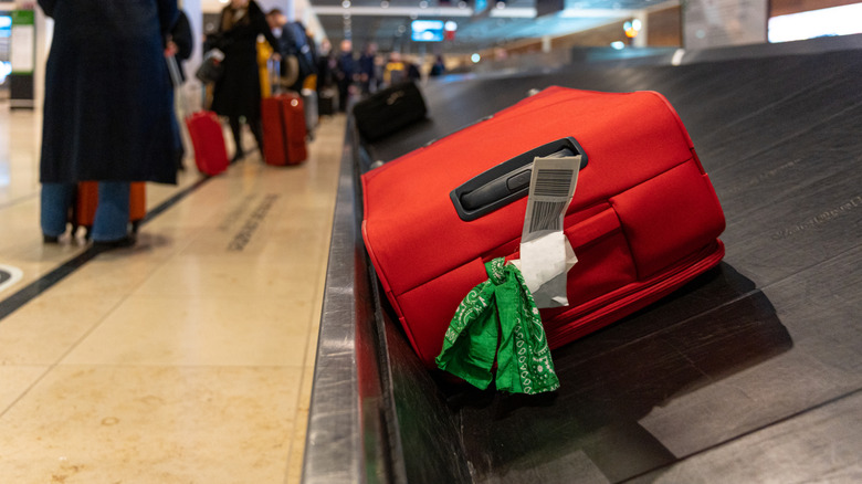 A red suitcase with a green bandana tied to the handle sits on a baggage claim carousel at an airport.