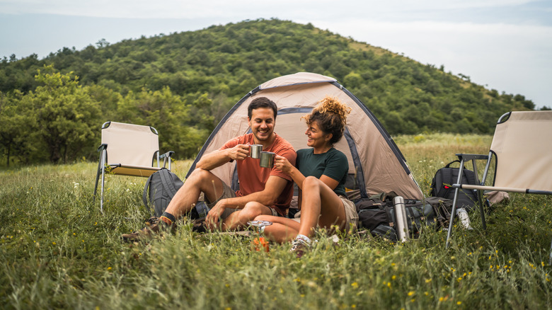 A man and woman enjoy camping, holding cups while sitting outside a tent