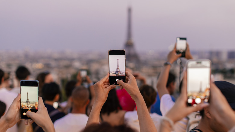 Crowds in Paris point their phones at the Eiffel Tower, waiting for the monument to light up at dusk.