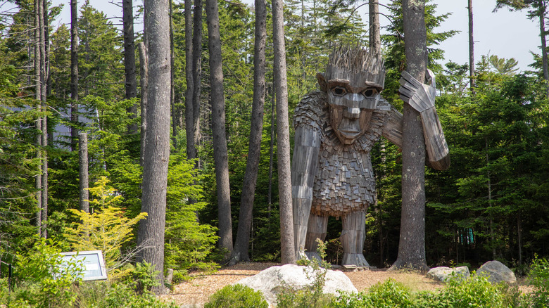A massive wood-sculpted troll stands among the trees of a forest