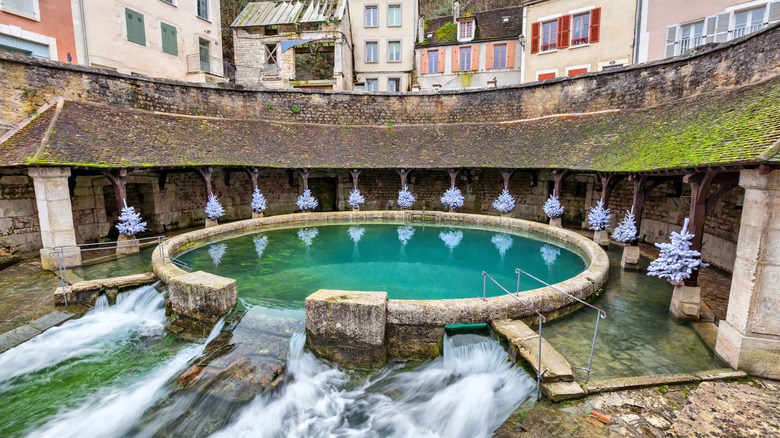 Round pool with turquoise water cascading into nearby stream surrounded by old buildings in a French building