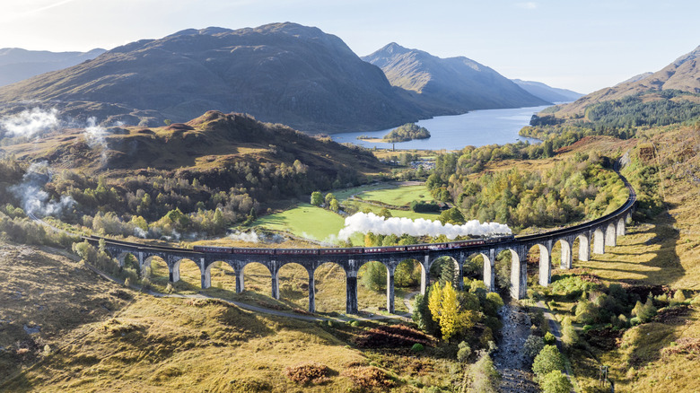 Scotland's red Jacobite Steam Train passing over the scenic Glenfinnan Viaduct near a lake, meadows, and mountains