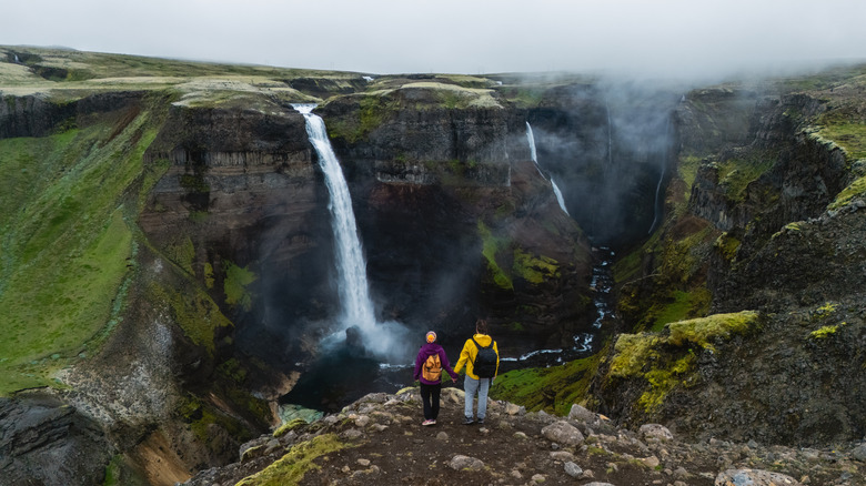 Two hikers stand at the edge of a dramatic cliff, gazing at the powerful Haifoss waterfall in Iceland