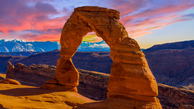 Utah's Delicate Arch rock formation standing tall in the mountains overlooking the snowy Rockies at sunset